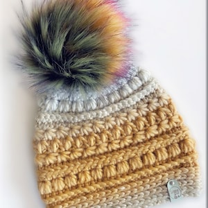 Könnte beinhalten: Handgehäkelte Mütze mit einem großen, flauschigen Kunstfell-Bommel. Die Mütze hat einen Farbverlauf, der von hellbeige zu goldgelb übergeht. Ein kleines Etikett mit dem Text "Loopy Suz" ist angebracht.