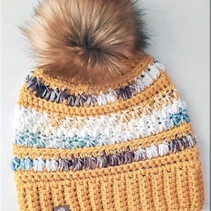 Puede incluir: Un gorro de punto con un pompón de piel sintética en color mostaza, con rayas blancas, marrones y azules. El gorro tiene un pequeño parche de cuero marrón con un corazón.
