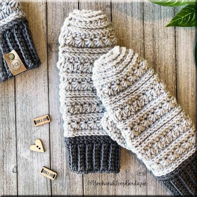 K&ouml;nnte beinhalten: Ein Paar graue und schwarze H&auml;kelhandschuhe mit einem strukturierten Stichmuster. Die Handschuhe haben einen grauen Bund und werden auf einem Holzgrund gezeigt.