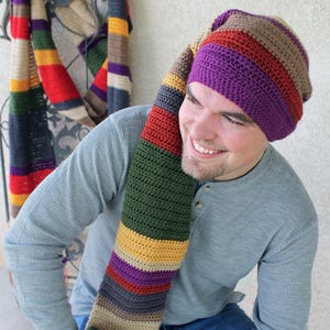 May include: A person wearing a long, striped, crocheted scarf and hat. The scarf is in shades of red, green, yellow, purple, brown, and gray. The hat is purple with brown, red, and gray stripes. The person is wearing a gray long-sleeved shirt and blue jeans.