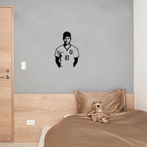 K&ouml;nnte beinhalten: Eine schwarz-wei&szlig;e Silhouette eines Fu&szlig;ballspielers, der ein Trikot mit der Nummer 10 tr&auml;gt. Der Spieler steht an einer grauen Wand mit einem Holz-Kopfteil und einer braunen Bettdecke dahinter.