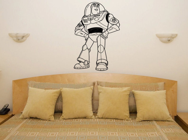 Puede incluir: Pegatina de pared de arte de l&iacute;nea en blanco y negro de Buzz Lightyear de la franquicia de pel&iacute;culas Toy Story. La pegatina est&aacute; en una pared blanca sobre una cama con ropa de cama de color canela.
