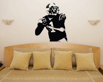 Op de afbeelding: Zwart-witte muursticker van een American footballspeler in een worphouding. De speler draagt een helm en een shirt met nummer 1. De sticker is geplaatst boven een bed met gele kussens.