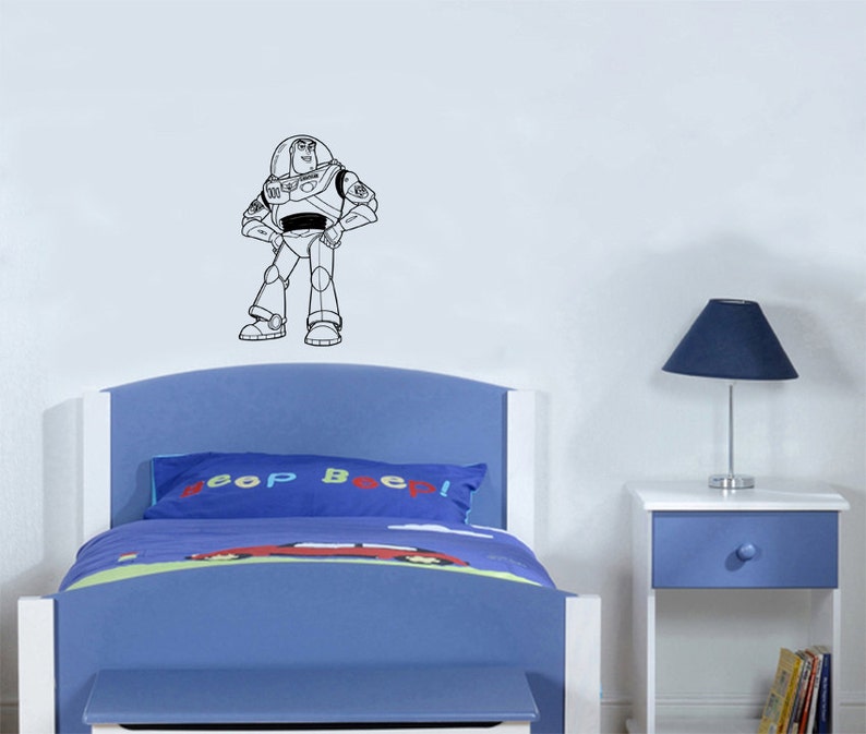 Puede incluir: Un contorno en blanco y negro de Buzz Lightyear de la serie de pel&iacute;culas Toy Story en una pared blanca sobre una cama azul con una almohada que dice "&iexcl;Bip Bip!"