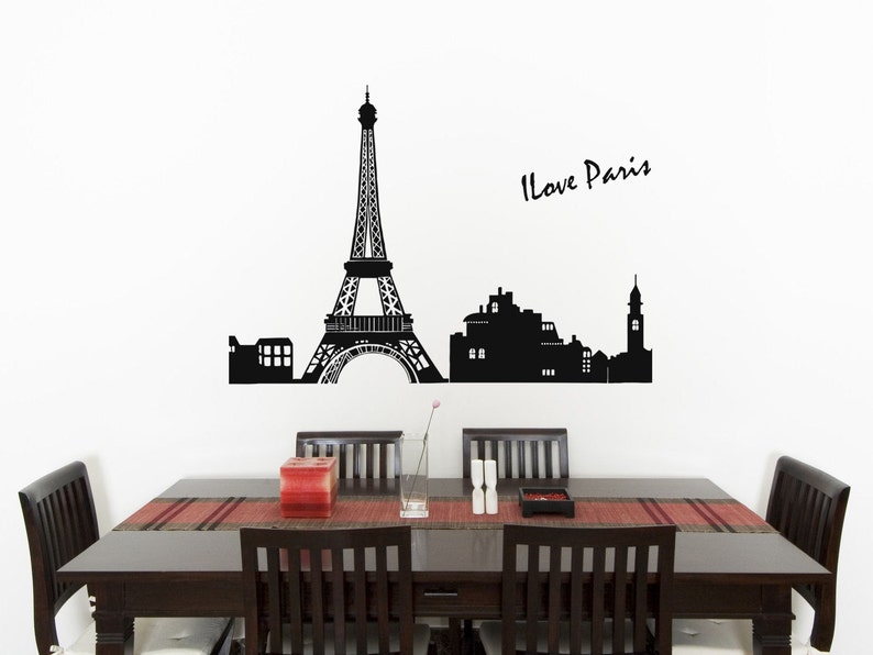 May include: Black and white silhouette wall decal of the Eiffel Tower and Parisian cityscape with the text "I Love Paris". The decal is displayed on a white wall above a dark wood dining table with six chairs.