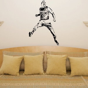Puede incluir: Pegatina de pared en silueta negra y blanca de un jugador de f&uacute;tbol en el aire, celebrando un gol. La pegatina se aplica a una pared sobre una cama con una cabecera de color marr&oacute;n y ropa de cama beige.