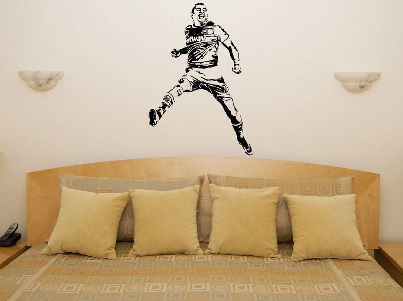 Puede incluir: Pegatina de pared en silueta negra y blanca de un jugador de f&uacute;tbol en el aire, celebrando un gol. La pegatina se aplica a una pared sobre una cama con una cabecera de color marr&oacute;n y ropa de cama beige.