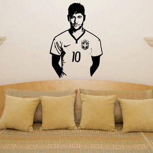 K&ouml;nnte beinhalten: Schwarze und wei&szlig;e Silhouette Wandaufkleber eines Fu&szlig;ballspielers, der ein Trikot mit der Nummer 10 tr&auml;gt. Der Aufkleber befindet sich an einer wei&szlig;en Wand &uuml;ber einem Bett mit beigen Kissen und einer gemusterten Bettdecke.