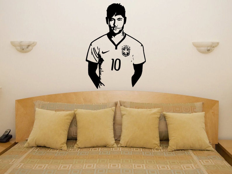 K&ouml;nnte beinhalten: Schwarze und wei&szlig;e Silhouette Wandaufkleber eines Fu&szlig;ballspielers, der ein Trikot mit der Nummer 10 tr&auml;gt. Der Aufkleber befindet sich an einer wei&szlig;en Wand &uuml;ber einem Bett mit beigen Kissen und einer gemusterten Bettdecke.