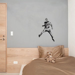 Puede incluir: Una silueta en blanco y negro de un jugador de f&uacute;tbol en el aire, pateando un bal&oacute;n. El jugador lleva una camiseta y pantalones cortos. La imagen est&aacute; en una pared gris sobre una cama con una cabecera marr&oacute;n y un edred&oacute;n marr&oacute;n. Un tigre de peluche est&aacute; sentado en la cama.