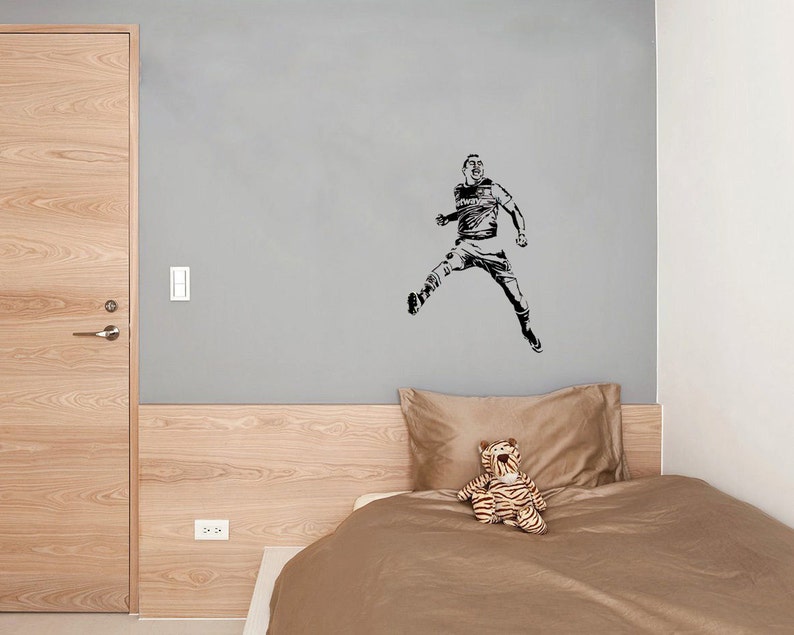 Puede incluir: Una silueta en blanco y negro de un jugador de f&uacute;tbol en el aire, pateando un bal&oacute;n. El jugador lleva una camiseta y pantalones cortos. La imagen est&aacute; en una pared gris sobre una cama con una cabecera marr&oacute;n y un edred&oacute;n marr&oacute;n. Un tigre de peluche est&aacute; sentado en la cama.