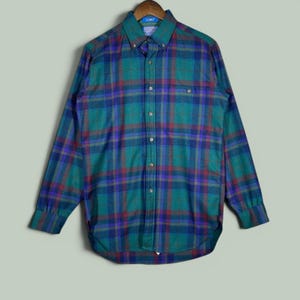 May include: A long-sleeve button-down shirt featuring a plaid pattern in shades of teal, dark blue, and deep red. The shirt has a pointed collar, a single chest pocket on the left side, and a full-length front button closure with matching buttons.