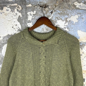 May include: An olive green knit cardigan featuring a ribbed collar, long sleeves, and a vertical cable knit pattern running down the center front. The garment is fastened with a single small button at the neckline and hangs on a wooden hanger.