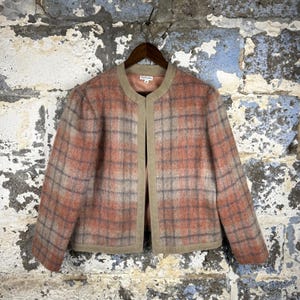 May include: A vintage, open-front jacket with a plaid pattern in shades of peach, gray, and beige. The jacket has a rounded neckline and is trimmed with a solid beige fabric. It is hanging on a wooden hanger against a textured wall.