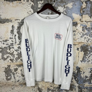 May include: A white long-sleeve shirt with the Bud Light logo on the chest and the word "BUD LIGHT" printed vertically on both sleeves in navy blue. The shirt is hanging on a wooden hanger.