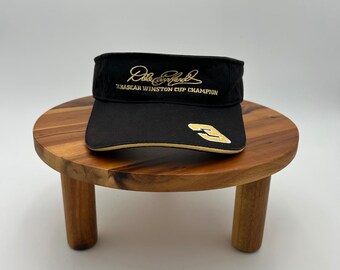 May include: A black visor featuring gold embroidery. The front displays the signature of Dale Earnhardt above the text NASCAR WINSTON CUP CHAMPION. A gold-outlined number 3 is embroidered on the right side of the brim with gold trim.