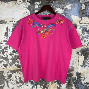 May include: A bright pink short-sleeved t-shirt with colorful embroidered floral designs on the front and around the neckline. The shirt is hanging on a wooden hanger against a textured wall. The brand name "LB DIFFUSION" is visible on the neckline.