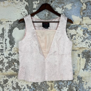 May include: A sleeveless pale pink vest featuring a subtle tonal floral jacquard pattern, a deep V-neckline, and a smooth interior lining. The garment includes a black brand label with the text ASHRO printed in red, hanging on a wooden hanger.