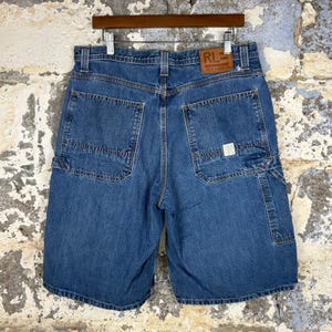 May include: Blue denim shorts featuring a brown leather patch on the back waistband with the text RL POLO JEANS CO. The shorts include two large back pockets, belt loops, and a small white rectangular label on the right back pocket.