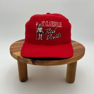 May include: A bright red baseball cap featuring an embroidered graphic of a devil figure holding a pitchfork. The text "ST. CLAIRSVILLE" is stitched in white above the words "Red Devils" in a stylized white font on the front panel.