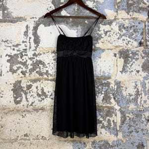 May include: A black knee-length dress featuring thin spaghetti straps, a square neckline, and a fitted bodice with a horizontal satin ribbon bow at the waist. The skirt is made of a sheer, layered fabric that falls in a straight, flowing silhouette.