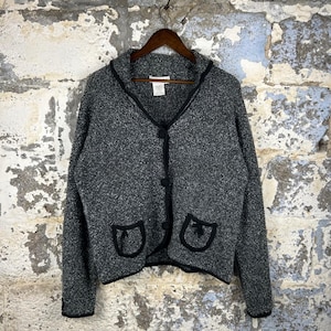 May include: A gray cardigan with black trim and buttons hangs on a wooden hanger. The sweater has two front pockets with black trim. The collar and edges of the cardigan are also trimmed in black.
