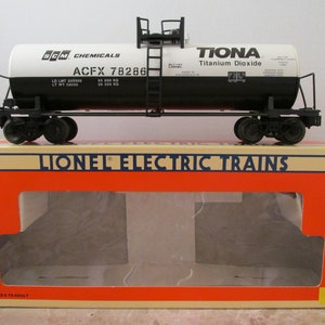 May include: A white and black Lionel Electric Trains model train car with a black tank car labeled "TIONA Titanium Dioxide". The car has a ladder on the side and is sitting on a brown cardboard box with the words "Lionel Electric Trains" printed on the front.