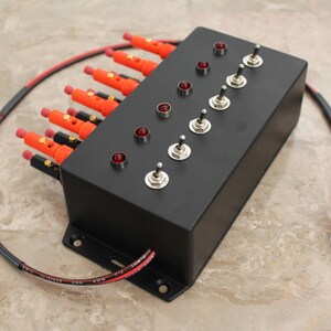 May include: A black box with eight toggle switches and red indicator lights. Each switch has a corresponding red LED light. The box has a black wire with red and black wires coming out of it.
