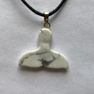 May include: A white marble whale tail pendant with black veining hangs from a black cord. The pendant is attached to a gold-colored bail. The necklace is displayed against a white background.