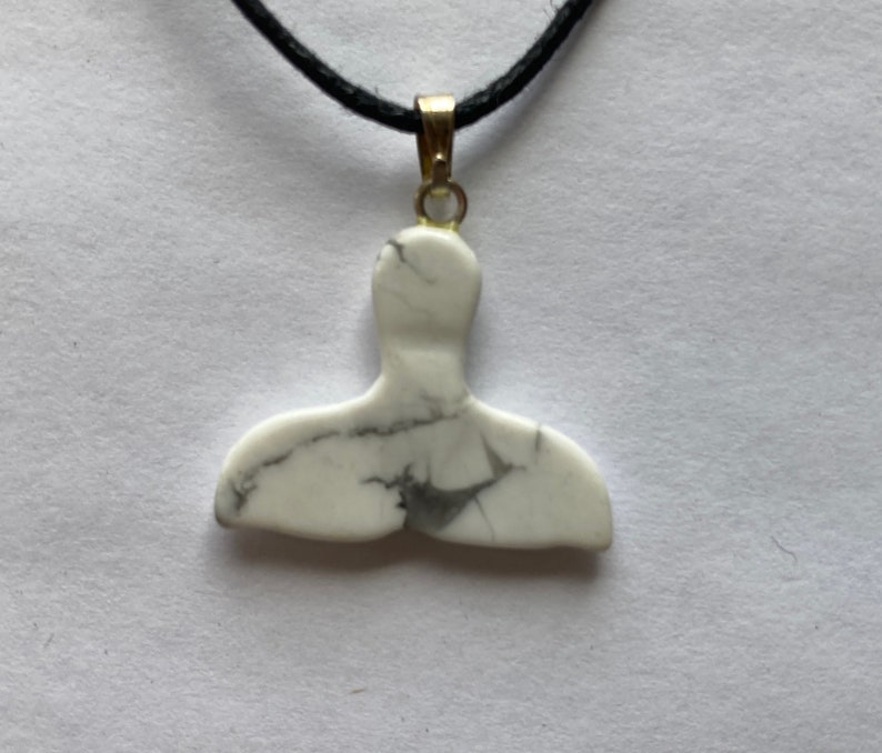 May include: A white marble whale tail pendant with black veining hangs from a black cord. The pendant is attached to a gold-colored bail. The necklace is displayed against a white background.