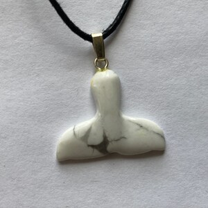 May include: A white and gray whale tail pendant on a black cord. The pendant is made of a marbled stone and has a gold-colored bail. The whale tail design is a popular nautical theme.