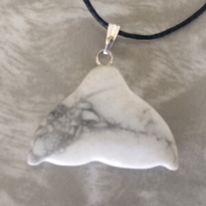 May include: A white stone pendant in the shape of a whale's tail, featuring grey veining. It hangs from a black cord with a silver-coloured clasp. This pendant is a distinctive piece of jewellery.