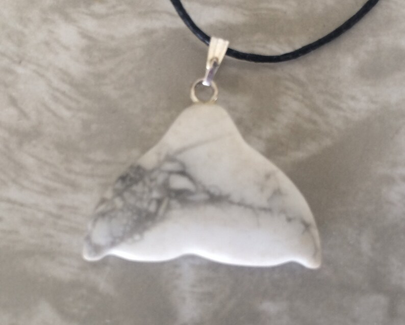 May include: A white stone pendant in the shape of a whale's tail, featuring grey veining. It hangs from a black cord with a silver-coloured clasp. This pendant is a distinctive piece of jewellery.