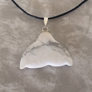 May include: White stone whale tail pendant with grey veining, suspended from a black cord necklace with a silver-coloured bail. The pendant's natural, organic shape is prominent. A simple, elegant piece of jewellery.