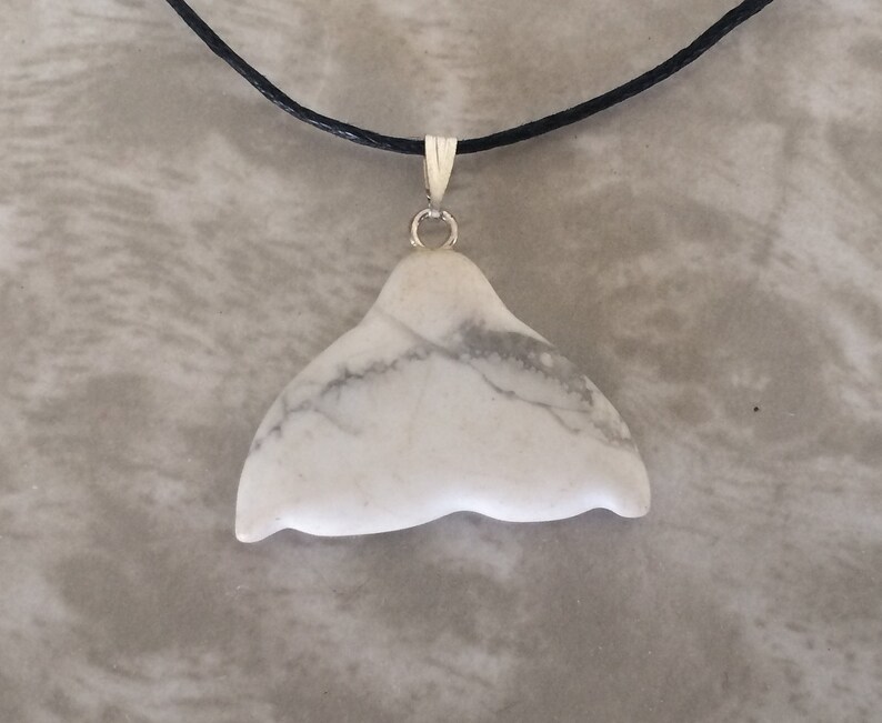 May include: White stone whale tail pendant with grey veining, suspended from a black cord necklace with a silver-coloured bail. The pendant's natural, organic shape is prominent. A simple, elegant piece of jewellery.