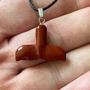 Op de afbeelding: Een rode edelsteen gegraveerd in de vorm van een walvisstaart, bevestigd aan een zwarte koordsnoer met een zilveren hanger.