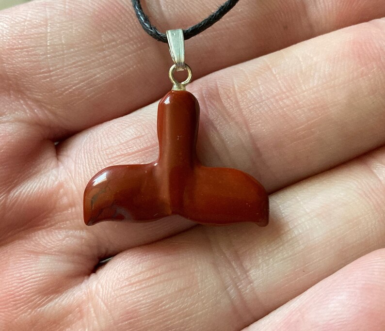 Op de afbeelding: Een rode edelsteen gegraveerd in de vorm van een walvisstaart, bevestigd aan een zwarte koordsnoer met een zilveren hanger.