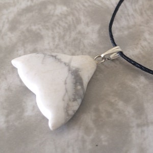May include: White howlite pendant with grey veining, shaped like a stylized bird or abstract form. The pendant is attached to a black cord necklace with a silver-coloured clasp. The pendant is a natural stone.