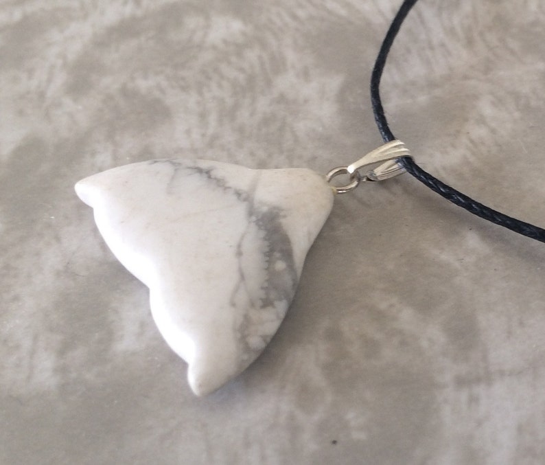 May include: White howlite pendant with grey veining, shaped like a stylized bird or abstract form. The pendant is attached to a black cord necklace with a silver-coloured clasp. The pendant is a natural stone.