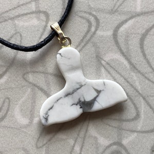 May include: White and gray marble pendant in the shape of a whale tail, suspended from a black cord necklace. The pendant is attached to a gold-colored bail. The necklace is displayed on a patterned surface.