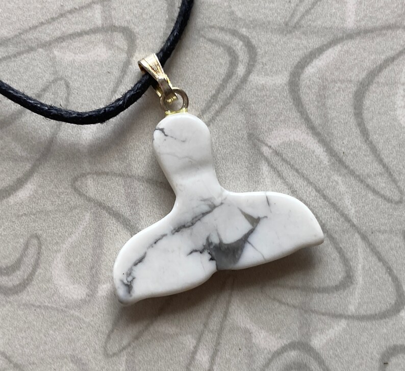 May include: White and gray marble pendant in the shape of a whale tail, suspended from a black cord necklace. The pendant is attached to a gold-colored bail. The necklace is displayed on a patterned surface.