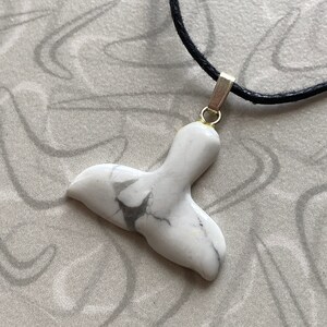 May include: A white marble pendant in the shape of a whale tail, with gray veining. The pendant is attached to a black cord necklace with a gold-colored bail.
