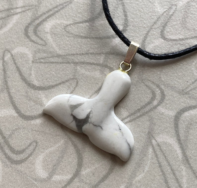 May include: A white marble pendant in the shape of a whale tail, with gray veining. The pendant is attached to a black cord necklace with a gold-colored bail.