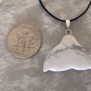 May include: A white marble whale tail pendant with grey veining, suspended from a black cord. A UK ten pence coin is next to the pendant for size comparison.