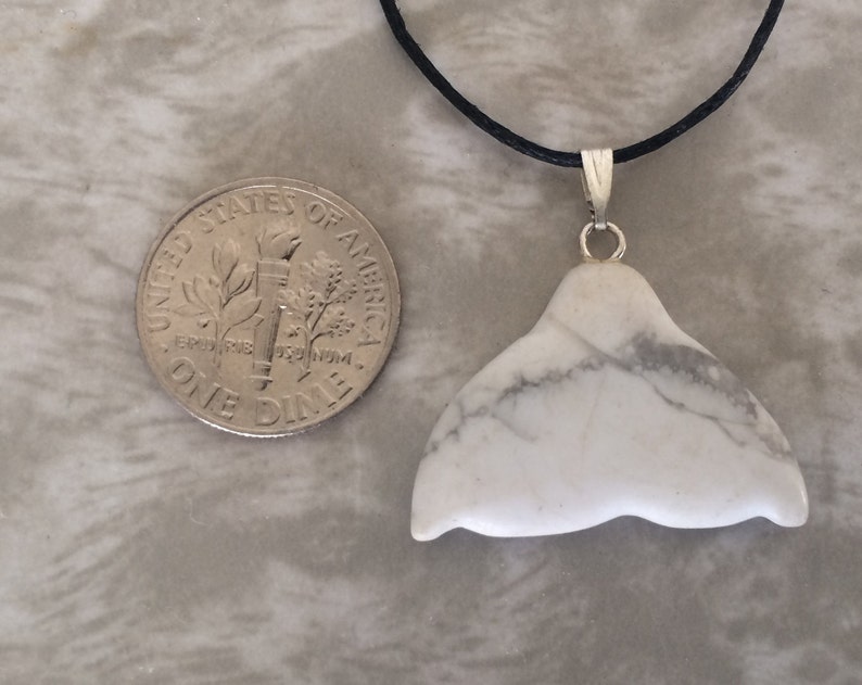 May include: A white marble whale tail pendant with grey veining, suspended from a black cord. A UK ten pence coin is next to the pendant for size comparison.
