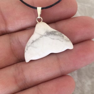 May include: A white whale tail pendant with grey marbling, suspended from a silver-coloured bail on a black cord. The pendant is made of a smooth, polished stone. The jewellery is held in a person's hand.
