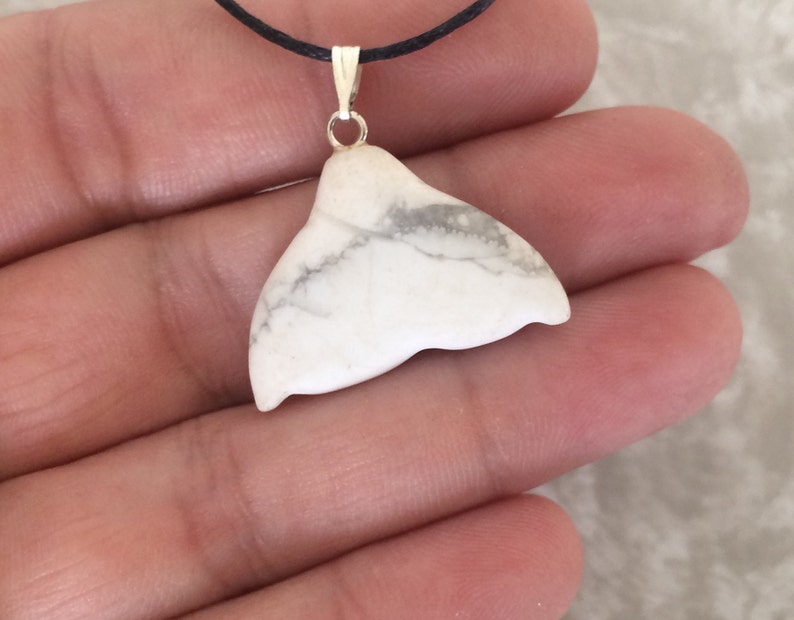 May include: A white whale tail pendant with grey marbling, suspended from a silver-coloured bail on a black cord. The pendant is made of a smooth, polished stone. The jewellery is held in a person's hand.