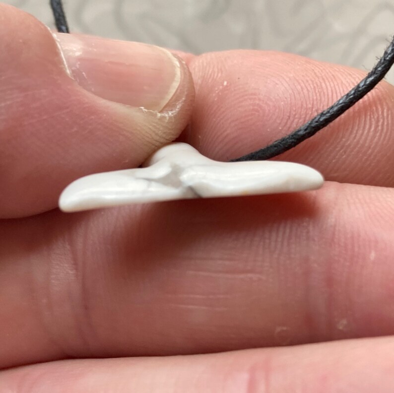 May include: A white pendant with gray veining, shaped like a whale tail, hangs from a black cord. The pendant is held between fingers, showcasing its smooth, polished surface. The jewelry is a minimalist design.
