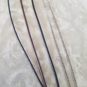May include: Close-up of several necklace chains and cords. The image shows a variety of materials, including black and brown cords, and silver-coloured chain necklaces. The cords have a woven texture, while the chains have a classic curb link design.