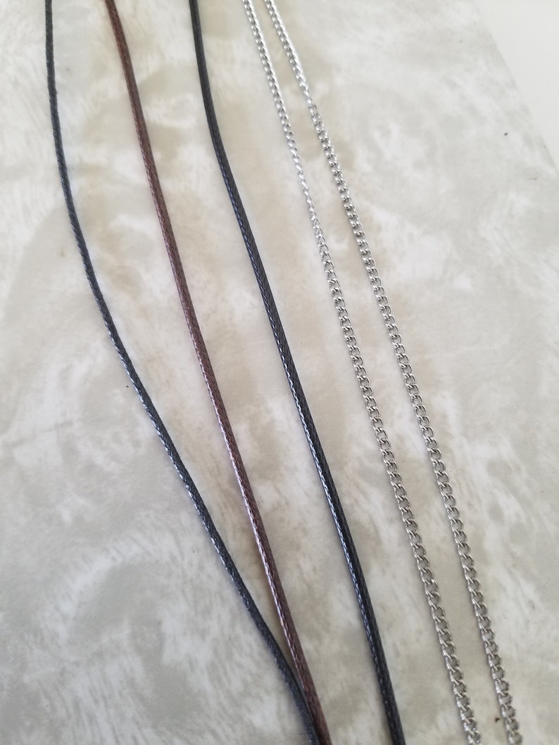 May include: Close-up of several necklace chains and cords. The image shows a variety of materials, including black and brown cords, and silver-coloured chain necklaces. The cords have a woven texture, while the chains have a classic curb link design.
